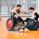 A player in a green and gold jersey grips the ball tightly while an opponent in a gray jersey attempts to block him. Both athletes are in motion, their sports wheelchairs colliding on the polished court.