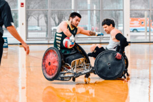 A player in a green and gold jersey grips the ball tightly while an opponent in a gray jersey attempts to block him. Both athletes are in motion, their sports wheelchairs colliding on the polished court.