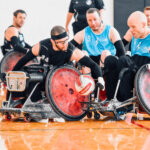 Two teams battle for possession of the ball in a wheelchair rugby match. A player in a black jersey (#24) leans forward with determination as he tries to block an opponent in a blue jersey (#15), who is maneuvering the ball with one hand.