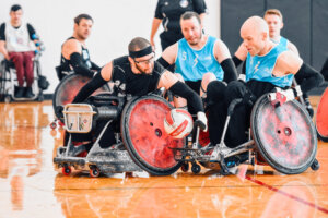 Two teams battle for possession of the ball in a wheelchair rugby match. A player in a black jersey (#24) leans forward with determination as he tries to block an opponent in a blue jersey (#15), who is maneuvering the ball with one hand.